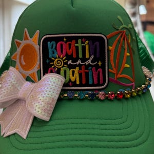 May include: Green trucker hat with a white sequined bow and a colorful embroidered patch that says "Boatin' and Datin". The hat also features a sun and sailboat design.
