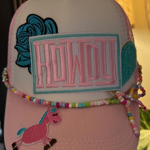 Puede incluir: Gorra de camionero con visera rosa y parte trasera de malla blanca. La gorra presenta un parche de caballo rosa y azul, una rosa azul y un parche que dice "HOWDY" en rosa. Una hebra de cuentas está envuelta alrededor de la visera.