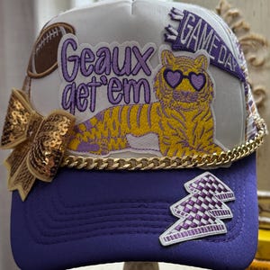 Ladies “Geaux Get ‘Em” Tiger trucker hat .    White and purple with football printed mesh .   Gold chain , rhinestones and gold sequin bow .