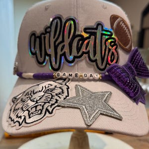 May include: A light-coloured baseball cap featuring a holographic 'wildcats' script, a brown football patch, a purple sequined bow, a beaded 'GAME DAY' strand, a white tiger head patch, and a silver glitter star patch on the front panel and brim.