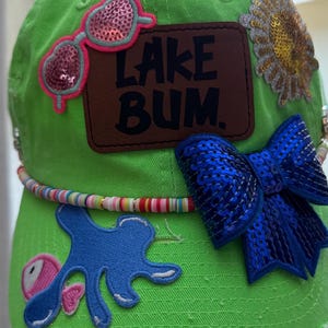 May include: Vibrant green baseball cap adorned with various patches. Features include sequined sunglasses, a sunflower, a blue sequined bow, and a brown patch with the text "LAKE BUM." A beaded necklace encircles the cap.