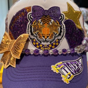 Puede incluir: Una gorra trucker blanca y morada con un parche central de tigre con ojos en forma de estrella y un lazo, acentuada por un gran lazo de lentejuelas doradas, un parche de estrella dorada, un corazón morado, cuentas moradas y el texto TIGERS.