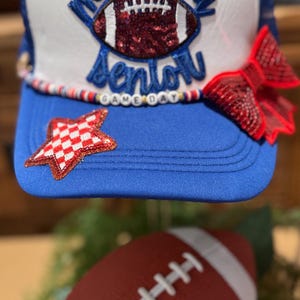 Pode incluir: Um boné de caminhoneiro azul e branco com o texto "Mom of a Senior" e um design de futebol americano. O chapéu tem um laço vermelho e um patch em forma de estrela. Uma bola de futebol americano vermelha e branca está em primeiro plano. "Game Day" está escrito no chapéu.