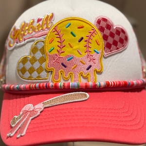 Puede incluir: Gorra de softbol blanca con visera rosa y una banda de cuentas coloridas. La gorra presenta parches bordados de una pelota de softbol, corazones y un bate con un lazo. La palabra "Softball" está bordada en la gorra.