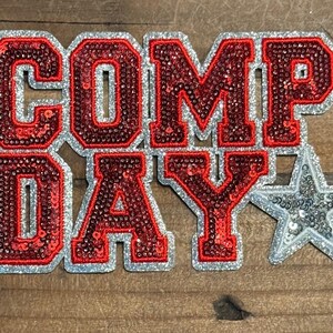 May include: A decorative patch featuring the text COMP DAY in bold, red, sequined varsity-style letters with a silver glitter border. A silver sequined star is positioned to the right of the word DAY, completing the design of this applique.