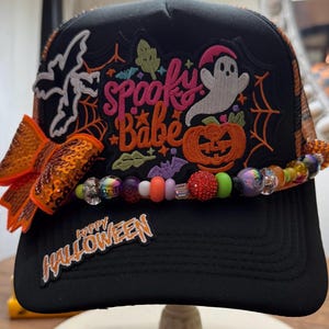 Könnte beinhalten: Eine schwarze Trucker-Cap mit einem großen gestickten Aufnäher mit dem Text Spooky Babe, einem weißen Geist, einem orangefarbenen Kürbis und einer Fledermaus. Die Kappe hat eine orangefarbene Pailletten-Schleife, bunte Perlen und den Text Happy Halloween.