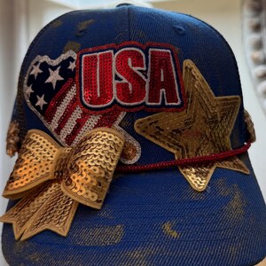 Könnte beinhalten: Blaue Baseballkappe mit goldenen Pailletten-Akzenten und patriotischem USA-Thema. Mit einer Pailletten-Schleife, einem Stern und einem Herz mit dem Design der amerikanischen Flagge. Das Wort "USA" ist aus roten Pailletten.