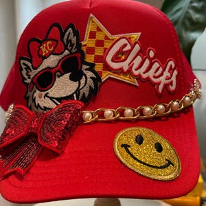 May include: A bright red trucker hat featuring a patch of a wolf wearing sunglasses and a bow, a yellow and red checkered star patch with the text Chiefs, a gold chain with white pearls, a large red sequined bow, and a gold glitter smiley face.