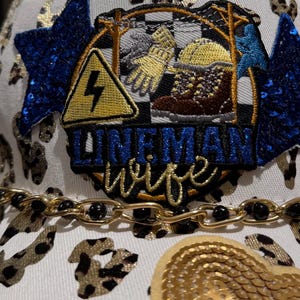 Può includere: Cappellino da baseball bianco con catena dorata e dettagli animalier. Il cappellino presenta una toppa con un fulmine giallo, scarponi da lavoro, guanti e la scritta "LINEMAN wife". Stelle di paillettes blu aggiungono un tocco decorativo.
