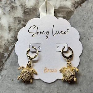 May include: Gold-colored brass earrings featuring a turtle design. The earrings are attached to a white card with the text "Shiny Luxe" in gold script. The word "Brass" is printed in gold.