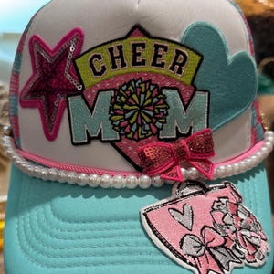 Puede incluir: Gorra de camionero turquesa y blanca con las palabras "CHEER MOM" en un diseño colorido. La gorra presenta una estrella de lentejuelas rosa, un corazón turquesa y un lazo rosa. Una hilera de perlas adorna el borde.