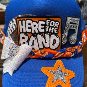 Puede incluir: Gorra de camionero azul real con el texto "HERE FOR THE BAND" en naranja y blanco. La gorra presenta acentos de fútbol americano, nota musical, estrella y lazo. El lazo dice "GAME DAY".