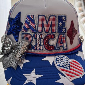 Könnte beinhalten: Eine weiß-blaue Trucker-Cap mit dem Schriftzug AMERICA in großen, texturierten Buchstaben. Das Design umfasst eine silberne Pailletten-Schleife, einen blauen Pailletten-Stern, eine rote Pailletten-Raute und einen herzförmigen Aufnäher.