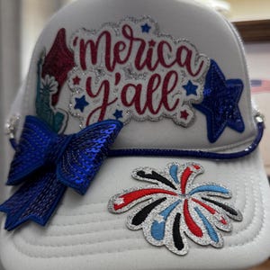 Może przedstawiać: Biała czapka typu trucker z czerwonym brokatowym napisem 'Merica y'all' i srebrnym brokatowym obrysem. Czapka ozdobiona jest niebieską kokardą z cekinów, niebieską gwiazdką z cekinów, naszywką Statuy Wolności oraz naszywką z motywem fajerwerków.