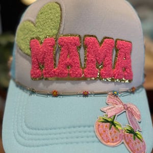 May include: A light blue trucker hat with a white mesh back. The hat has a pink fuzzy "MAMA" patch with gold glitter trim, a green fuzzy heart patch, and a pink and green cherry patch with a pink bow.