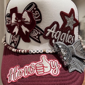 Puede incluir: Una gorra trucker blanca y granate con un parche en forma de Texas con un lazo blanco, una estrella plateada, un lazo de lentejuelas plateadas y un parche que dice Aggies. Un cordón de cuentas dice GAME DAY sobre un parche de Howdy.