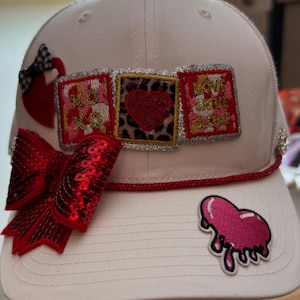 Puede incluir: Gorra de béisbol blanca con adornos rojos y rosas. Cuenta con un gran lazo rojo con lentejuelas, un pequeño lazo a cuadros blanco y negro y parches en forma de corazón. La gorra tiene tres parches cuadrados con diseños de corazón y estampado de leopardo.