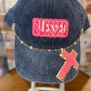 Może przedstawiać: Czapka bejsbolowa z ciemnoniebieskiego denimu z naszywką „Blessed” w kolorze różowym i różowym krzyżem z ozdobnym złotym brokatem. Czapka ma ozdobny pasek z koralikami i kolorowymi kwiatami.