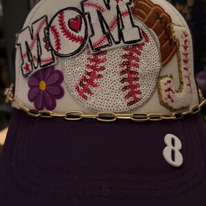 Puede incluir: Gorra de béisbol blanca con ribete morado y una cadena dorada. La gorra tiene varios parches con temática de béisbol, incluyendo una pelota de béisbol grande con lentejuelas, un guante y un parche que dice "MOM" con un corazón rojo. El número "8" está en la parte posterior de la gorra.