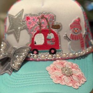 Op de afbeelding: Een turquoise en witte truckerpet met feestelijke versieringen. Het heeft een zilveren paillettenstrik, een zilveren ster en een rode auto met cadeaus. Een sneeuwpop en een roze sneeuwvlok met een juweel maken het ontwerp af.