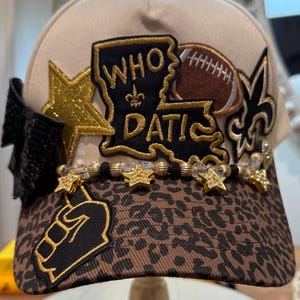 May include: A beige baseball cap with a leopard print brim, featuring a black patch shaped like Louisiana with the text WHO DAT! and a fleur-de-lis. It includes a brown football patch, a gold glitter star, a black bow, and a beaded star trim.