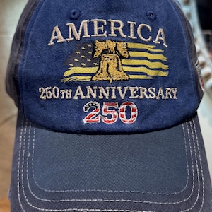 May include: A navy blue baseball cap with the word "AMERICA" embroidered in tan above a gold Liberty Bell and a stylized American flag. The text "250th ANNIVERSARY" and "250" are below the flag. The brim has the dates "1776-2026".