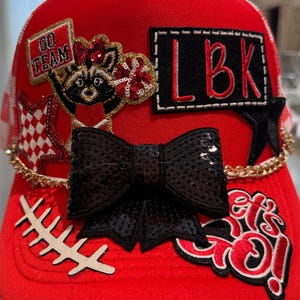 May include: A bright red trucker hat adorned with various patches including a raccoon cheerleader, a black bow with sequins, a gold chain, and text patches reading GO TEAM, LBK, and let's go. The hat features a white checkered star and a football lace.