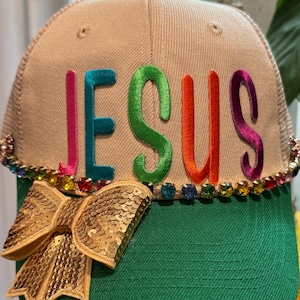 Puede incluir: Una gorra tipo camionero beige y verde con la palabra JESUS en letras bordadas de colores en la parte frontal. La gorra está decorada con una hilera de pedrería multicolor en la base de la corona y un gran lazo de lentejuelas doradas en el lateral.