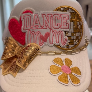 May include: A white trucker hat featuring a patch that reads DANCE MOM in pink and white lettering. The hat is decorated with a red heart, a gold disco ball, a gold sequined bow, a gold chain link trim, and a yellow flower patch with a pink center.