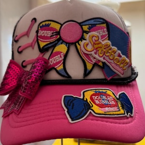 Könnte beinhalten: Rosa und weißer Trucker-Hut mit schwarzem Mesh-Rücken. Der Hut hat eine große Schleife und mehrere Aufnäher, darunter einen "Softball"-Aufnäher und einen "Doubles Bubble"-Bonbon-Aufnäher. Die Schleife ist gelb, rosa und blau.