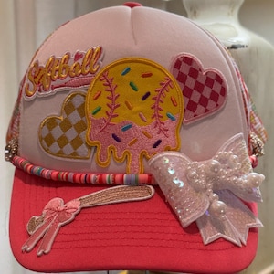 Puede incluir: Gorra de softbol rosa y blanca con visera roja. La gorra presenta un parche en forma de softbol con chispas, un lazo y la palabra "Softball". Corazones decorativos y un bate de béisbol completan el diseño.