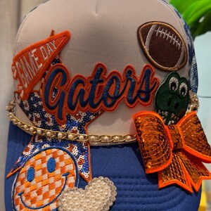 May include: A blue and white trucker hat featuring an orange pennant with the text GAME DAY, a brown football patch, a green alligator patch, a blue and orange checkered smiley face, a heart made of white pearls, and an orange sequin bow.