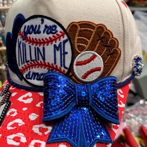 May include: Cream-colored baseball cap with a red and white leopard print brim. The cap features a blue sequined bow, a baseball glove, and a baseball patch with the text "you're killin me". A small blue jewel is on the side.