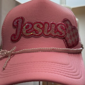 Puede incluir: Gorra de camionero rosa con la palabra "JESUS" en letras de lentejuelas y un corazón de lentejuelas con un patrón de cuadros. Una cadena de perlas rodea el sombrero, atada con un lazo. La gorra tiene una visera curva.