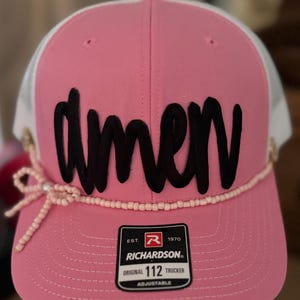Op de afbeelding: Roze en witte truckerpet met het woord "amen" in zwart script. De pet heeft een kralenband en een strikdetail. De Richardson 112 Trucker is verstelbaar.