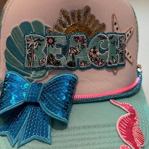 May include: A turquoise and white trucker hat with the word "BEACH" embroidered in turquoise, featuring a sequined bow, a seahorse, and a starfish. The hat has a pink and blue cord detail.