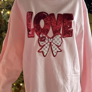 May include: A light pink sweatshirt with the word "LOVE" in red sequined letters. Below the word is a bow with a checkered pattern and a kiss design. The sweatshirt is displayed on a mannequin.