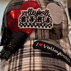 Puede incluir: Gorra de béisbol a cuadros grises y blancos, adornada con parches. Presenta un corazón rojo con lentejuelas, un parche con la palabra "volleyball" y gráficos de voleibol, y un parche que dice "I ❤️ Volleyball!"