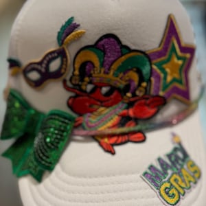 May include: White trucker hat with Mardi Gras patches. It features a red crawfish wearing a jester hat, a green sequined bow, a purple star, and a mask. The words "Mardi Gras" are also on the hat.
