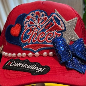 Puede incluir: Una gorra de camionero roja con un gran parche bordado con la palabra Cheer, un megáfono y un pompón. Incluye una estrella de purpurina plateada, un lazo de lentejuelas azules, una hilera de perlas blancas y un parche negro que dice I love Cheerleading!