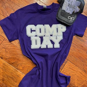 May include: Purple short-sleeved t-shirt with the words "COMP DAY" in white glitter letters.