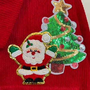 Pode incluir: Camisola de veludo cotelê vermelho com uma árvore de Natal de lantejoulas verdes e douradas e uma aplicação de Papai Noel vermelha e branca.