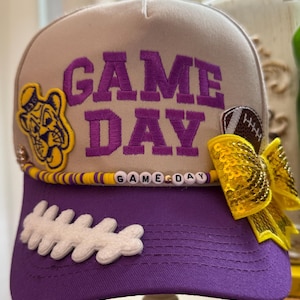 Damska fioletowo-złota czapka typu trucker GAME DAY. Koralikowy łańcuszek i kokarda z cekinów w kolorze żółtego złota. Liczne naszywki. Filcowa naszywka z motywem tygrysa w stylu retro.