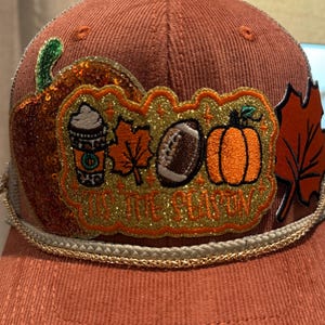 Peut inclure: Casquette de baseball couleur rouille avec une texture en velours côtelé. Le chapeau présente un écusson décoratif avec une tasse de café, un ballon de football américain et une citrouille, avec le texte "IT'S THE SEASON". Une bordure en corde dorée orne le bord.