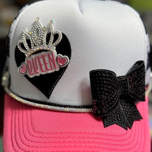 Puede incluir: Gorra de camionero con un panel frontal blanco y una visera rosa. El sombrero presenta un corazón negro con la palabra "QUEEN" en rosa, una corona enjoyada y un lazo negro con lentejuelas. Una banda de pedrería adorna la visera.