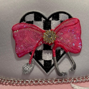 May include: A white hat with a pink and silver bow on a black and white chequered heart. The hat has a pink chain trim.