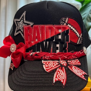 Könnte beinhalten: Eine schwarze Trucker-Cap mit einem rot-schwarzen Stickaufnäher mit der Aufschrift RAIDER POWER. Die Kappe ist mit einem Strass-Stern, einer roten Schleife mit Strass-Mitte, einem geflochtenen roten Bandana-Besatz und einer roten Paisley-Schleife verziert.