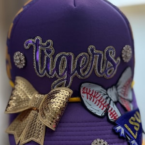 Puede incluir: Gorra de béisbol morada con la palabra "Tigers" en escritura brillante. La gorra presenta lazos de lentejuelas doradas, detalles de pedrería y un lazo con temática de béisbol. Una pequeña bandera de LSU está adjunta al costado.