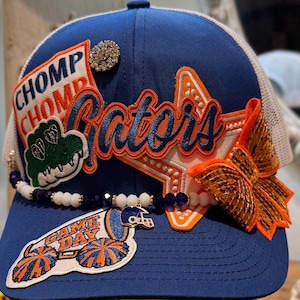May include: A blue baseball cap featuring a white mesh back, decorated with orange and blue patches that read CHOMP CHOMP and Gators, a green alligator patch, a GAME DAY megaphone patch, a sequined orange bow, and a beaded strand with a helmet charm.
