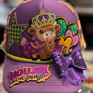 May include: A purple baseball cap featuring a cow wearing a crown and cape, with the text "Boujee on a budget". The cap has a sequined bow and star, with gold, green, and purple accents. The brim is checkered.